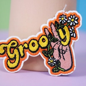 May include: An embroidered patch with the word "groovy" in yellow and orange lettering, with a pink hand holding a flower stem with white daisies.