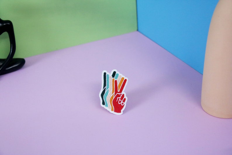 Pu&ograve; includere: Toppa ricamata colorata a forma di mano che fa il segno della pace. La mano &egrave; rossa con dita nei toni dell'arancione, giallo, verde acqua e nero. La toppa ha il testo "766" sulla mano rossa.
