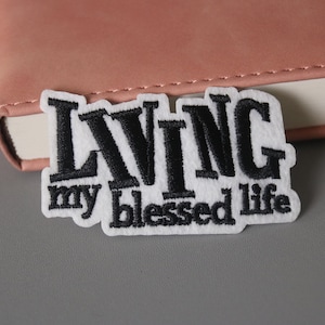 May include: White embroidered patch with black lettering that reads "LIVING my blessed life".