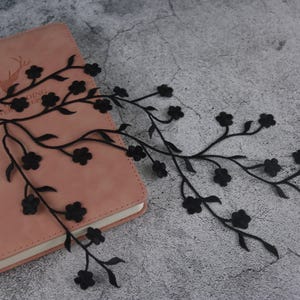 Puede incluir: Borde de encaje negro con un diseño floral. El borde presenta ramas delicadas con pequeñas flores negras. El borde se muestra en una libreta rosa.