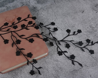 May include: Black lace trim with a floral design. The trim features delicate branches with small black flowers. The trim is displayed on a pink notebook.