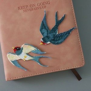 May include: Two embroidered swallow bird patches on a pink leather journal with the text "Keep on going never give up".