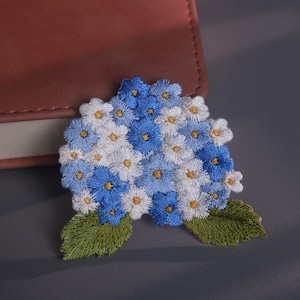 May include: Embroidered floral patch featuring a cluster of blue and white flowers with yellow centers and green leaves. The patch is placed on a gray surface next to a brown leather-bound book.