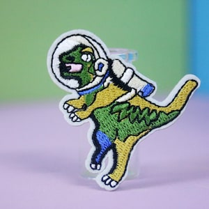 Pode incluir: Um remendo bordado de um dinossauro verde usando um capacete de astronauta branco com um design azul e amarelo. O dinossauro está usando um traje espacial amarelo com uma faixa azul.
