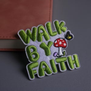 May include: Embroidered patch with the words "WALK BY FAITH" in green, featuring a red mushroom with white spots and a yellow butterfly. The patch is on a white background.