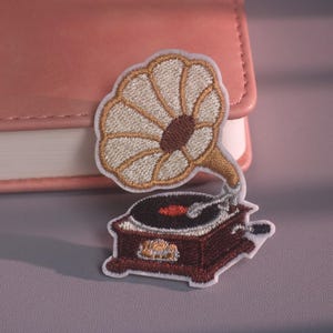Puede incluir: Un parche bordado que muestra un gramófono antiguo con una bocina grande de color claro, una base de color marrón oscuro y un disco negro. El diseño está delineado con hilo blanco, mostrando costuras detalladas en el interior de la bocina.