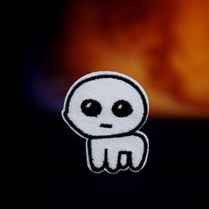May include: A white embroidered patch featuring a minimalist cartoon character with a round head, large black eyes, and a small mouth. The character has a simple body with four short legs. The patch is outlined in black.