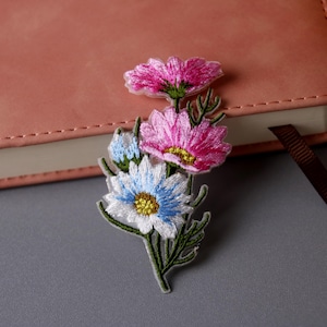 Op de afbeelding: Een set van drie geborduurde bloemenpatches in roze, wit en blauw. De patches bevinden zich op een grijs oppervlak.