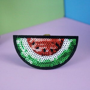 Può includere: Un cerotto a forma di fetta di anguria verde, bianca e rossa fatta di paillettes. Il cerotto è nero e ha un contorno nero.