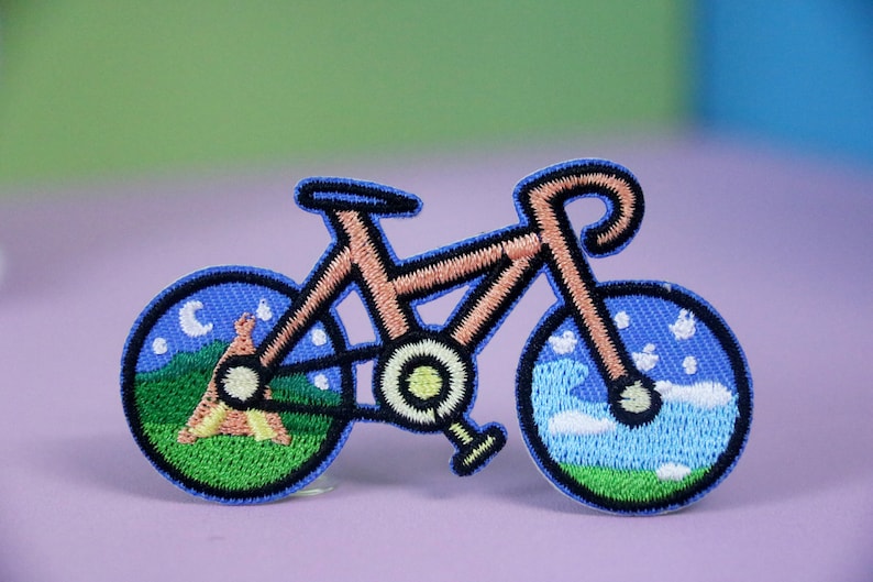 May include: An embroidered patch depicting a bicycle with a campfire scene in one wheel and a night sky scene in the other wheel.