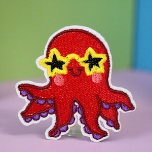 May include: Red embroidered octopus wearing yellow star-shaped sunglasses. The octopus has eight tentacles with purple accents.