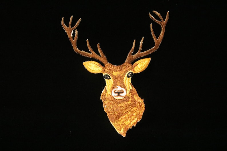 May include: Embroidered patch featuring a deer head with large antlers. The deer has a brown coat, yellow ears, and a white nose, set against a black background. This patch is suitable for apparel and accessories.