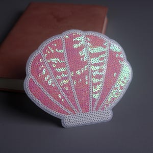 May include: A pink sequined seashell patch with a white border. The patch is on a grey surface with a brown leather-bound notebook in the background. The sequins shimmer, reflecting light.