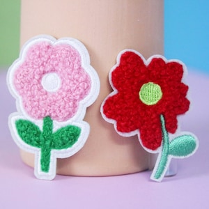 May include: Two embroidered felt flower patches. One is pink with a white center and green leaves. The other is red with a green center and green leaves.