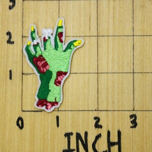May include: Green zombie hand with red and yellow details. The hand is raised with fingers outstretched. The hand is on a wooden surface with a grid pattern.