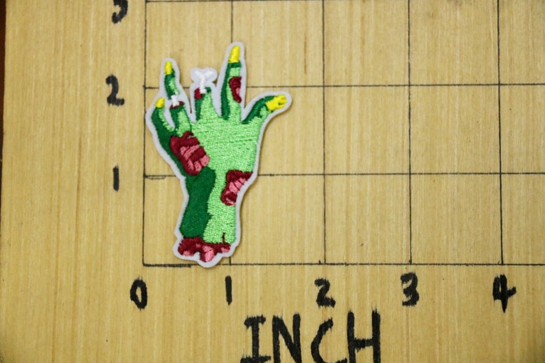 May include: Green zombie hand with red and yellow details. The hand is raised with fingers outstretched. The hand is on a wooden surface with a grid pattern.