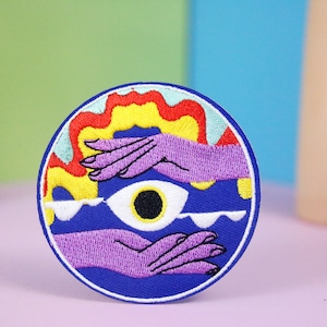 May include: Embroidered patch with a vibrant design. The patch features a blue circular background with a white border. The design includes a large eye, two purple hands, and colorful flames in yellow, red, and orange.