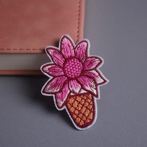 May include: Embroidered patch of a pink flower with white and pink detailing, set on a brown, textured base. The patch has a white border and is against a grey background, suggesting a floral theme.