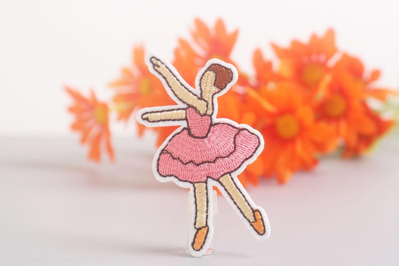 May include: Embroidered patch of a ballerina in a pink tutu, arms outstretched. The patch has a white border and is set against a backdrop of orange flowers. The ballerina's hair is brown, and her shoes are orange.