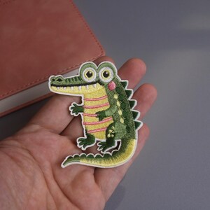 May include: Embroidered alligator patch with a cartoon design. The patch features a green body with yellow and pink accents, large eyes, and a white border. This patch is suitable for clothing and accessories.