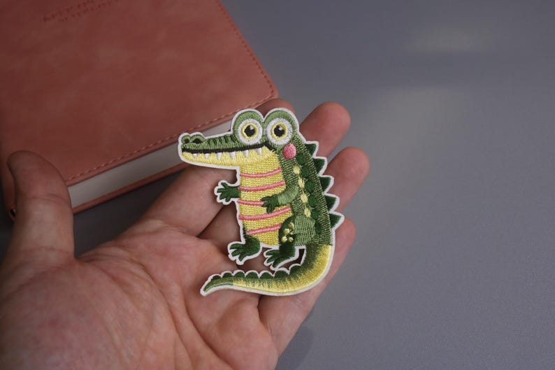May include: Embroidered alligator patch with a cartoon design. The patch features a green body with yellow and pink accents, large eyes, and a white border. This patch is suitable for clothing and accessories.