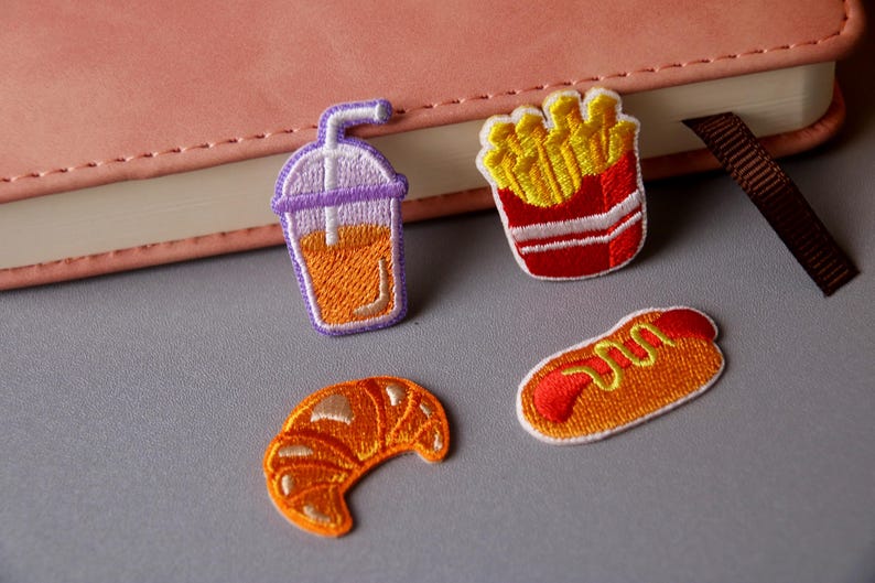 May include: Four embroidered iron-on patches featuring a croissant, a hot dog, french fries, and a glass of iced tea.