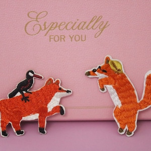 Może przedstawiać: Dwa haftowane naszywki z motywem lisów. Jeden lis nosi kapelusz i okulary przeciwsłoneczne, drugi ma ptaka siedzącego na grzbiecie.