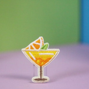 Puede incluir: Un parche bordado que representa un vaso de cóctel con una rodaja de naranja y una hoja verde en la parte superior.