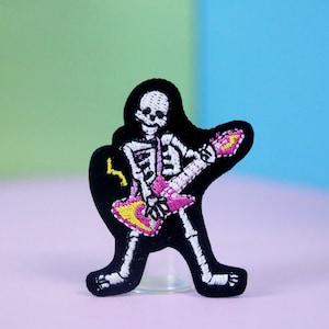 Könnte beinhalten: Schwarzer gestickter Aufnäher mit einem Skelett, das eine pinkfarbene E-Gitarre mit gelben Akzenten spielt.