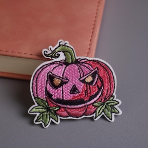 Könnte beinhalten: Gestickter Halloween-Patch mit einem lächelnden Kürbis in Rosa und Rot. Der Patch hat einen grünen Stiel und Blätter, vor einem neutralen Hintergrund. Ideal, um Kleidung oder Accessoires einen gruseligen Touch zu verleihen.