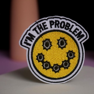 May include: Embroidered patch with a yellow smiley face design featuring bullet hole details. The patch has the text "I'M THE PROBLEM" in a black font, outlined in white. The patch is on a white background.