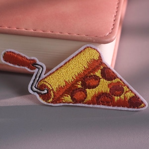 Puede incluir: Un parche bordado con forma de porción de pizza de pepperoni siendo aplicada por un rodillo de pintura. La porción de pizza presenta una corteza y queso amarillos con círculos de pepperoni rojo oscuro. El rodillo tiene un mango plateado y agarre naranja.