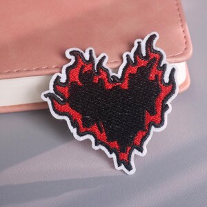 Puede incluir: Parche bordado con un diseño de corazón negro con llamas rojas, bordeado en blanco. El parche está sobre una superficie rosa claro. Un artículo decorativo para ropa o accesorios.