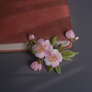 Peut inclure: Un journal relié en cuir rose avec une broderie florale. La broderie présente des fleurs roses avec des centres jaunes, des feuilles vertes et des tiges marron. Le journal est fermé et posé sur une surface grise.