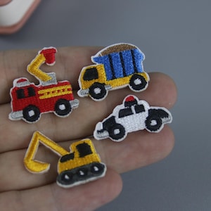 May include: Four embroidered iron-on patches featuring different vehicles: a red fire truck, a yellow dump truck, a white police car, and a yellow excavator.