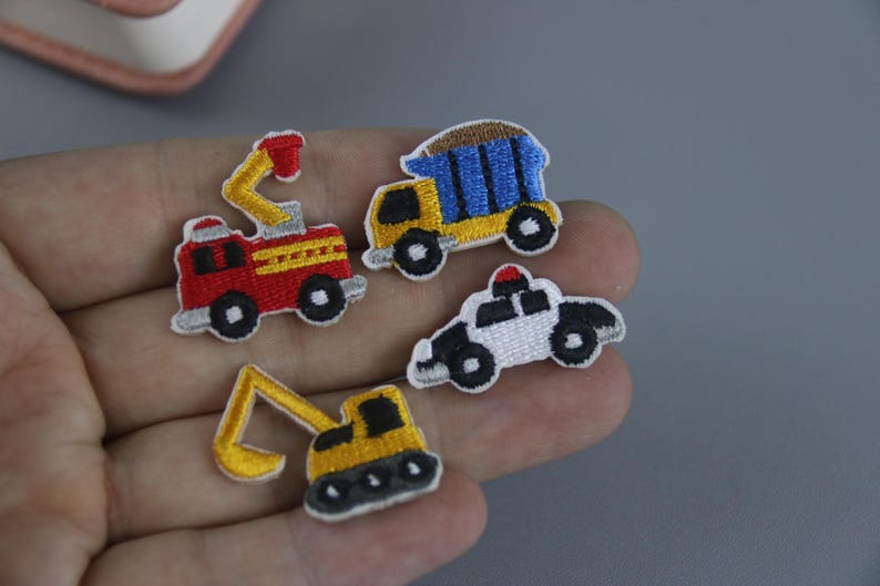 May include: Four embroidered iron-on patches featuring different vehicles: a red fire truck, a yellow dump truck, a white police car, and a yellow excavator.