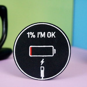Peut inclure: Un patch rond noir avec une broderie blanche. Le patch représente une icône de batterie avec une barre rouge indiquant 1% de charge. Le texte "1% I'M OK" est au-dessus de l'icône de batterie.