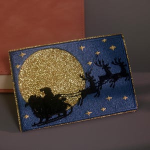 May include: A rectangular card featuring a dark blue background with a large, circular, gold glitter moon. A black silhouette of Santa Claus in a sleigh pulled by reindeer flies across the moon, surrounded by small gold glitter stars.