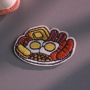 Può includere: Una toppa ricamata con il disegno di una colazione inglese completa. La toppa raffigura due uova al tegamino, strisce di pancetta, salsicce, una fetta di pane tostato, una fetta di pomodoro e fagioli, contornati da filo nero su fondo bianco.