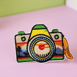 Puede incluir: Un parche bordado de colores para planchar que representa una c&aacute;mara con una escena de puesta de sol en el visor. La c&aacute;mara es amarilla, verde, azul y roja. El parche tiene una correa de cuero marr&oacute;n.