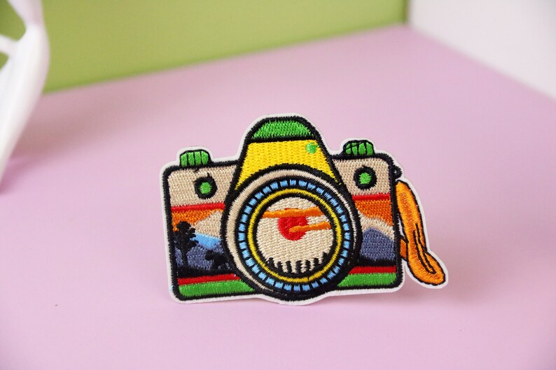 Puede incluir: Un parche bordado de colores para planchar que representa una c&aacute;mara con una escena de puesta de sol en el visor. La c&aacute;mara es amarilla, verde, azul y roja. El parche tiene una correa de cuero marr&oacute;n.