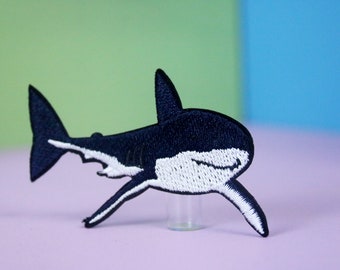May include: A black and white embroidered shark patch. The shark is facing right and has a white underbelly.