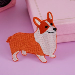 Peut inclure: Patch brodé représentant un chien corgi brun et blanc avec un visage souriant. Le patch est sur un fond violet clair.