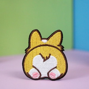Puede incluir: Parche bordado de la parte trasera de un corgi marrón y amarillo con una cola blanca en forma de corazón. El corgi está sentado con las patas metidas debajo del cuerpo y tiene almohadillas rosadas.