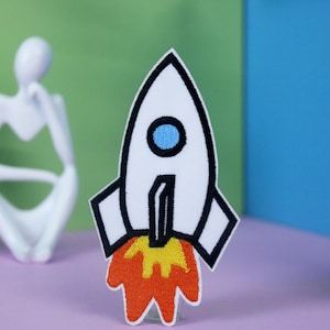 May include: White rocket ship with black outline and a blue window. The rocket is on a pink surface and has orange flames coming out of the bottom.