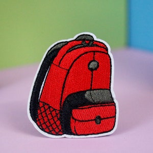 May include: Red embroidered iron-on patch of a backpack with a black zipper and a black pocket.