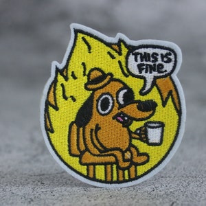 May include: A yellow and orange embroidered patch featuring a cartoon dog sitting in a chair with a cup of coffee. The dog is wearing a hat and has a speech bubble above its head that says "This is fine."