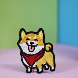 Pode incluir: Etiqueta bordada de um cão Shiba Inu usando uma bandana vermelha. O cão está sorrindo com a língua para fora. A etiqueta é amarela, branca e vermelha.