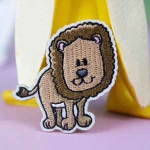 May include: A brown embroidered lion patch with a white outline. The lion has a pink nose and pink inside its ears. The lion is standing on all fours and has a small tail.