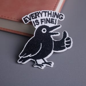 May include: Embroidered patch featuring a black crow with a white outline, giving a thumbs-up. The text "EVERYTHING IS FINE!" arches above the bird. The patch is on a gray surface, with a pink notebook visible in the background.